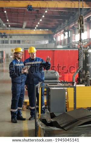 Two engineers standing in large workshop or plant and discussing new industrial equipment or machine while one of them pointing at it