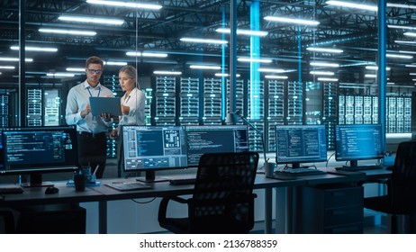 Two IT Engineers Standing in Working Data Center. They Use Laptop while Standing at the Evening Office. Team of Developers Look at Each Other, Talk and Using Computer - Powered by Shutterstock