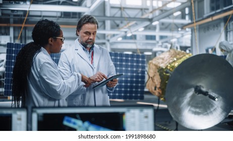 Two Engineers Talk, Use Tablet Computer while Working on Satellite Construction. Aerospace Agency Manufacturing Facility: Scientists Build, Assemble Spacecraft for Space Exploration Mission
