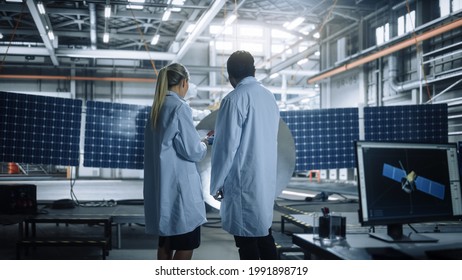 Two Engineers Talk, Use Tablet Computer while Working on Satellite Construction. Aerospace Agency Manufacturing Facility: Scientists Build, Assemble Spacecraft for Space Exploration Mission