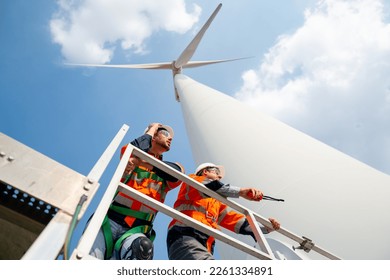 Two engineers or technician workers stand on base of big windmill or wind turbine and look to right side with blue sky.