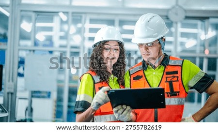 Two engineers use a tablet to program an AI system. They are in a futuristic smart factory, reviewing data for industrial robots or microchip manufacturing.