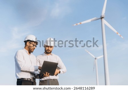 Two engineers using a laptop are analyzing the operation of wind turbines in a wind farm, promoting sustainable energy and environmental protection