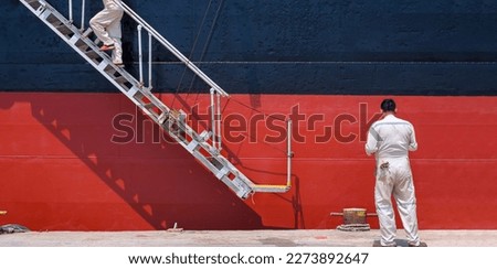 Two Engineers are Walking up the white gangway Accommodation Ladder of red and black Oil Tanker to work on the Ship at Shipyard