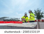 Two engineers wearing high visibility jackets discuss construction progress near concrete barricades on-site. One holds a laptop, symbolizing modern planning and collaboration in civil engineering.