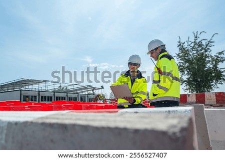 Two engineers wearing high visibility jackets discuss construction progress near concrete barricades on-site. One holds a laptop, symbolizing modern planning and collaboration in civil engineering.