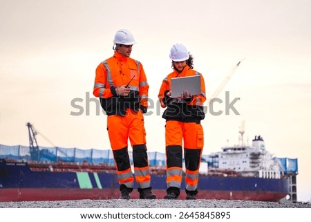 Two engineers wearing high-visibility safety gear and hard hats review plans on a laptop at a shipyard, with a large cargo ship and cranes in the background during sunset.