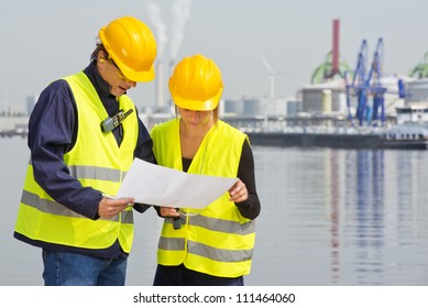 Two Engineers, Wearing Safety Gear, Including Goggles, Hard Hat, Ear Plugs And Reflective Safety Vests, Discussing Blue Prints In An Industrial Harbor With Factories And Plants In The Background