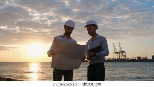 Two Engineers In White Safety Helmets Work With A Blueprint On A Paper Drawing Against The Backdrop Of The Seaport In The Early Morning.