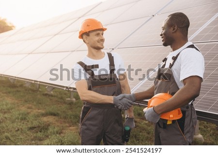 Two engineers in work overalls shake hands at a solar panel installation site, symbolizing teamwork and renewable energy. They wear protective helmets on a sunny day.