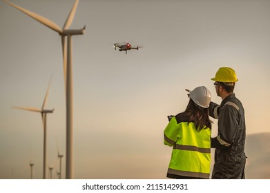 Two engineers working and holding the report at wind turbine farm Power Generator Station on mountain,Thailand people,Technician man and woman discuss about work,Use drone view from high angle