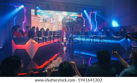 Two Esport Teams of Pro Gamers Play in FPS Shooter Video Game on a Championship Arena with Big Screen Showing Mock-up Gameplay. Cyber Games Tournament Event with Competing Players, Spectators Cheer