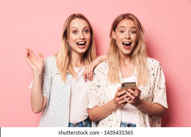 Two excited attractive blonde girls wearing summer outfit standing isolated over pink background, using mobile phone