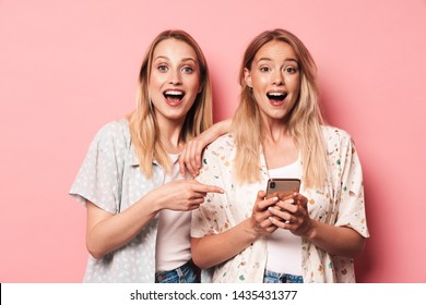 Two excited attractive blonde girls wearing summer outfit standing isolated over pink background, pointing finger at mobile phone