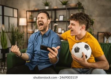 Two excited caucasian young adult men friends or brother use phone to watch football match and cheer at home - Powered by Shutterstock