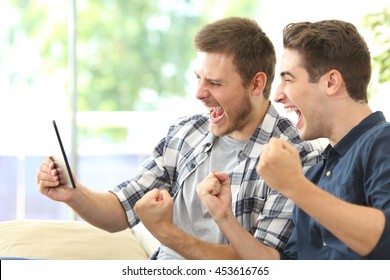 Two excited friends or roommates watching tv on line in a tablet sitting on a couch in the living room at home