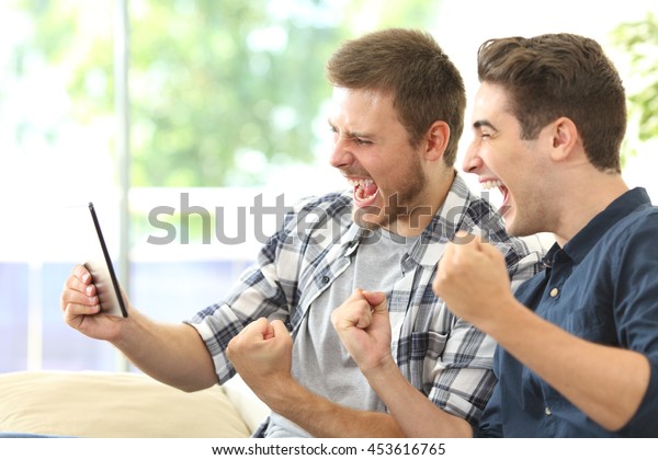 Two excited\
friends or roommates watching tv on line in a tablet sitting on a\
couch in the living room at\
home