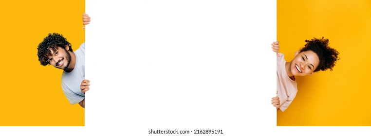 Two excited people, african american girl and indian or arabian guy, peeking out from blank white mockup banner, empty space for presentation, advertising, stand on isolated orange background, smile