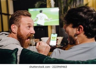 Two excited white men watching football match   making bets at bookmaker's website while sitting sofa in front TV screen