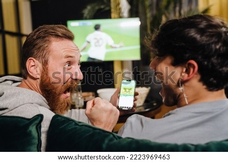 Two excited white men watching football match and making bets at bookmaker's website while sitting on sofa in front of TV screen