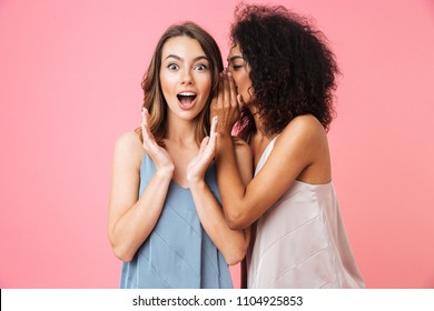 Two excited young girls dressed in summer clothes whispering secrets isolated over pink background