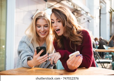 Two excited young girls using mobile phones while sitting at the cafe outdoors and pointing finger - Powered by Shutterstock