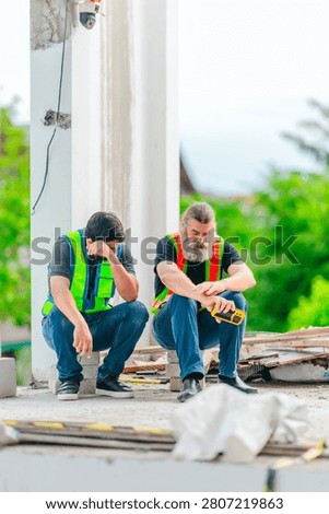 Two exhausted construction workers crouch and rest with burnout, tired and stressed after hard manual labor at a house building site outdoors.