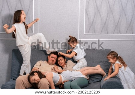 Two exhausted young parents are sitting on the couch while their four daughters are running around and screaming. Difficulties of a large family concept.