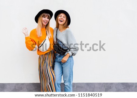 Two  exited hipster women posing over white wall. Fall season. Wearing stylish orange  knitted sweater, black hat , bum bag. Friends spending great time together . Copy space .