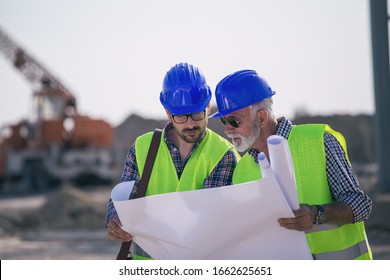 Two Experienced Engineers Looking At Blueprints And Talking At Building Site With Metal Crane In Background