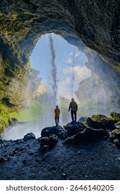 Two explorers stand before the breathtaking Kvernufoss waterfall in Iceland, mesmerized by the cascading water and brilliant landscape. A sunny day enhances the vibrant scenery.