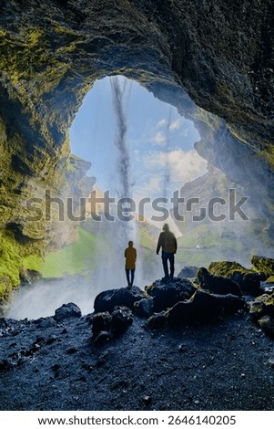 Two explorers stand before the breathtaking Kvernufoss waterfall in Iceland, mesmerized by the cascading water and brilliant landscape. A sunny day enhances the vibrant scenery.