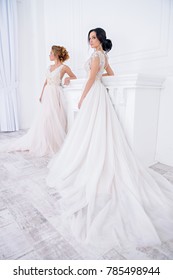 Two Exquisite Beautiful Brides In Elegant Wedding Dresses Stand In White Luxurious Apartments. Wedding Fashion. 