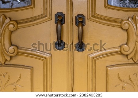 Two exterior vintage doors of a house with metal door knobs. The wooden decorative doors are yellow in color. There's an ornate or fancy style molding around two brass metal handles on the doors. 