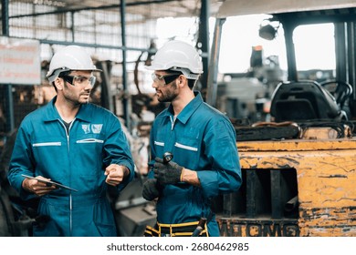 Two factory workers are collaborating on a large piece of industrial machinery. Both men are wearing blue jumpsuits, black work gloves, and white hard hats, with also wearing safety glasses.