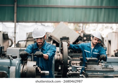 Two factory workers are collaborating on a large piece of industrial machinery. Both men are wearing blue jumpsuits, black work gloves, and white hard hats, with also wearing safety glasses.