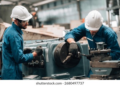 Two factory workers are collaborating on a large piece of industrial machinery. Both men are wearing blue jumpsuits, black work gloves, and white hard hats, with also wearing safety glasses.
