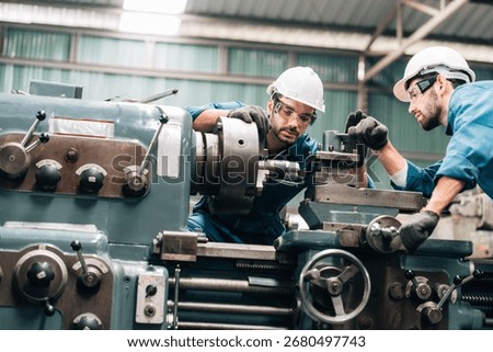 Two factory workers are collaborating on a large piece of industrial machinery. Both men are wearing blue jumpsuits, black work gloves, and white hard hats, with also wearing safety glasses.