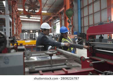 Two factory workers examine a machine in a workshop. They wear safety helmets and discuss the operation of the equipment. The setting has other machines in the background.