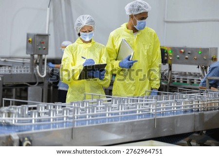 Two factory workers in protective uniforms and face masks inspect canned food production line with laptop in manufacturing. Food safety, quality control, hygiene, industrial inspection concept.