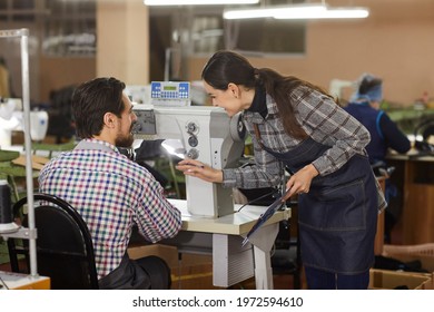 Two factory workers talking at table with industrial shoe sewing machine. Workshop manager discussing production process changes with coworker. Footwear manufacturing industry and equipment concept