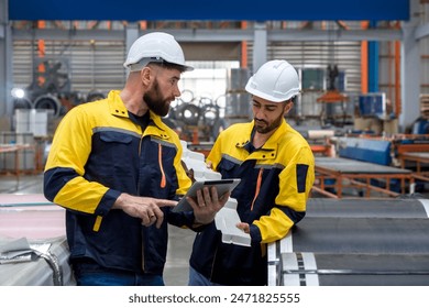 Two factory workers in work uniform and protective hardhat, inspecting a stack of large steel coils in an industrial setting or manufacturing facility. - Powered by Shutterstock