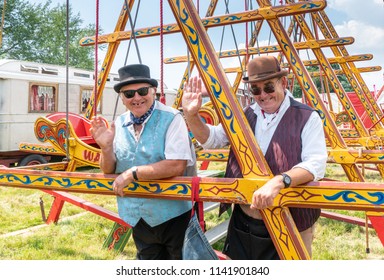 Two Fairground Workers With Bowler Hat, Neckerchief And Blue Or Mauve Waistcoat Smile Broadly And Wave. They Lean Against Vibrant Yellow Swing Boats In Front Of A Traditional Travelling Van.