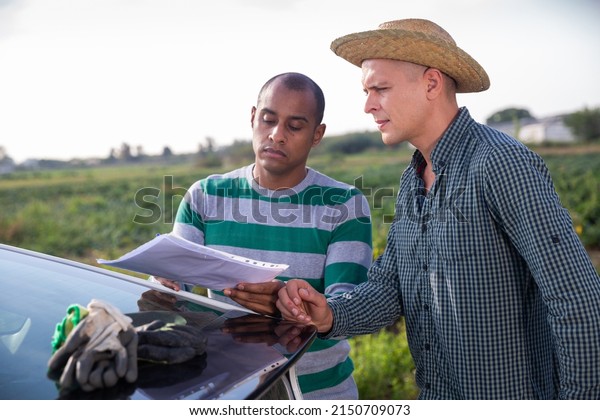 Two farmers discussing papers while standing\
near car on farm field on autumn\
day
