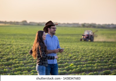 Two Farmers Man And Woman With Tablet Standing In Soybean Field In Front Of Tractor At Sunset