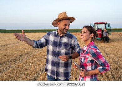 Two Farmers Man And Woman Talking On Wheat Field During Harvest With Tractor In Background In Summer Time