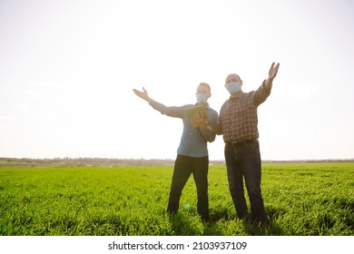 Two Farmers In Sterile Medical Masks Discuss Agricultural Issues On A A Green Wheat Field. Farmers With Tablet In The Field. Smart Farm. Agro Business. Covid-19.