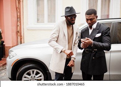 Two fashion black men stand near business car and look at cell phone. Fashionable portrait of african american male models. Wear suit, coat and hat.