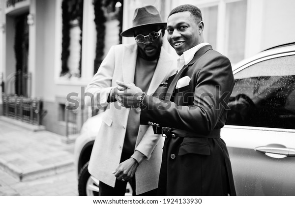 Two fashion black men stand near business\
car and look at cell phone. Fashionable portrait of african\
american male models. Wear suit, coat and\
hat.