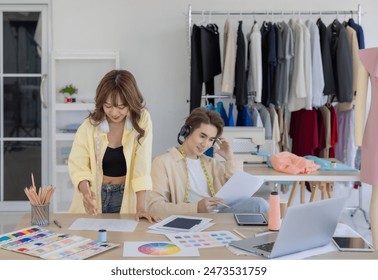 Two fashion designers enjoying creative workspace, surrounded by fabric samples, color palettes, clothing racks. One designer is listening to music with headphones while other is engrossed in tablet. - Powered by Shutterstock